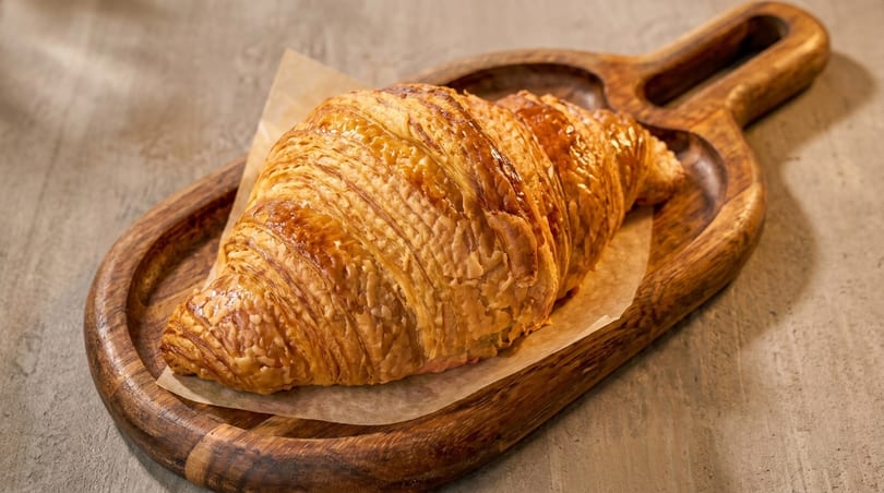 Golden-brown croissant on parchment paper, served on a rustic wooden tray.