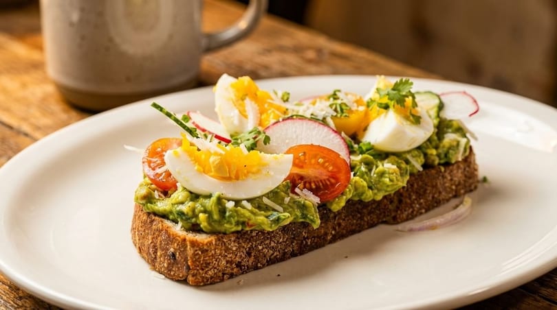Avocado toast topped with sliced hard-boiled eggs, cherry tomatoes, radishes, and cilantro on a white plate.