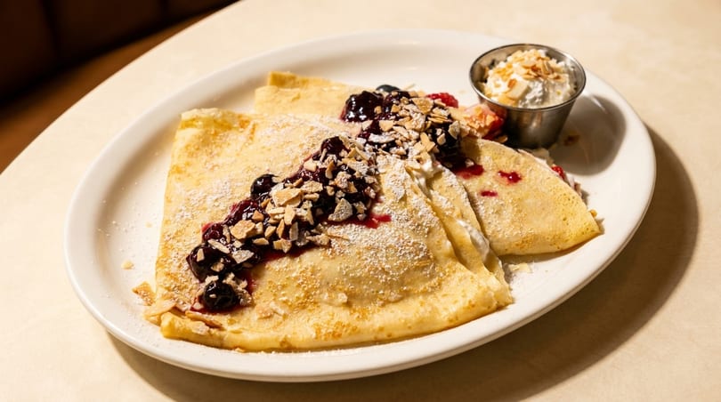 Three golden crepes topped with berry compote, sliced almonds, powdered sugar, and a side of whipped cream.