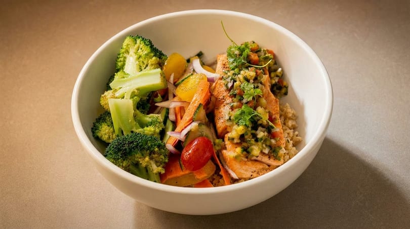 A white bowl filled with salmon, rice, broccoli, and mixed vegetables on a neutral background.