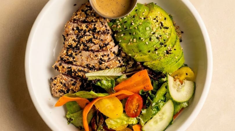Seared tuna with sesame seeds, sliced avocado, mixed greens, and a side of dressing in a white bowl.